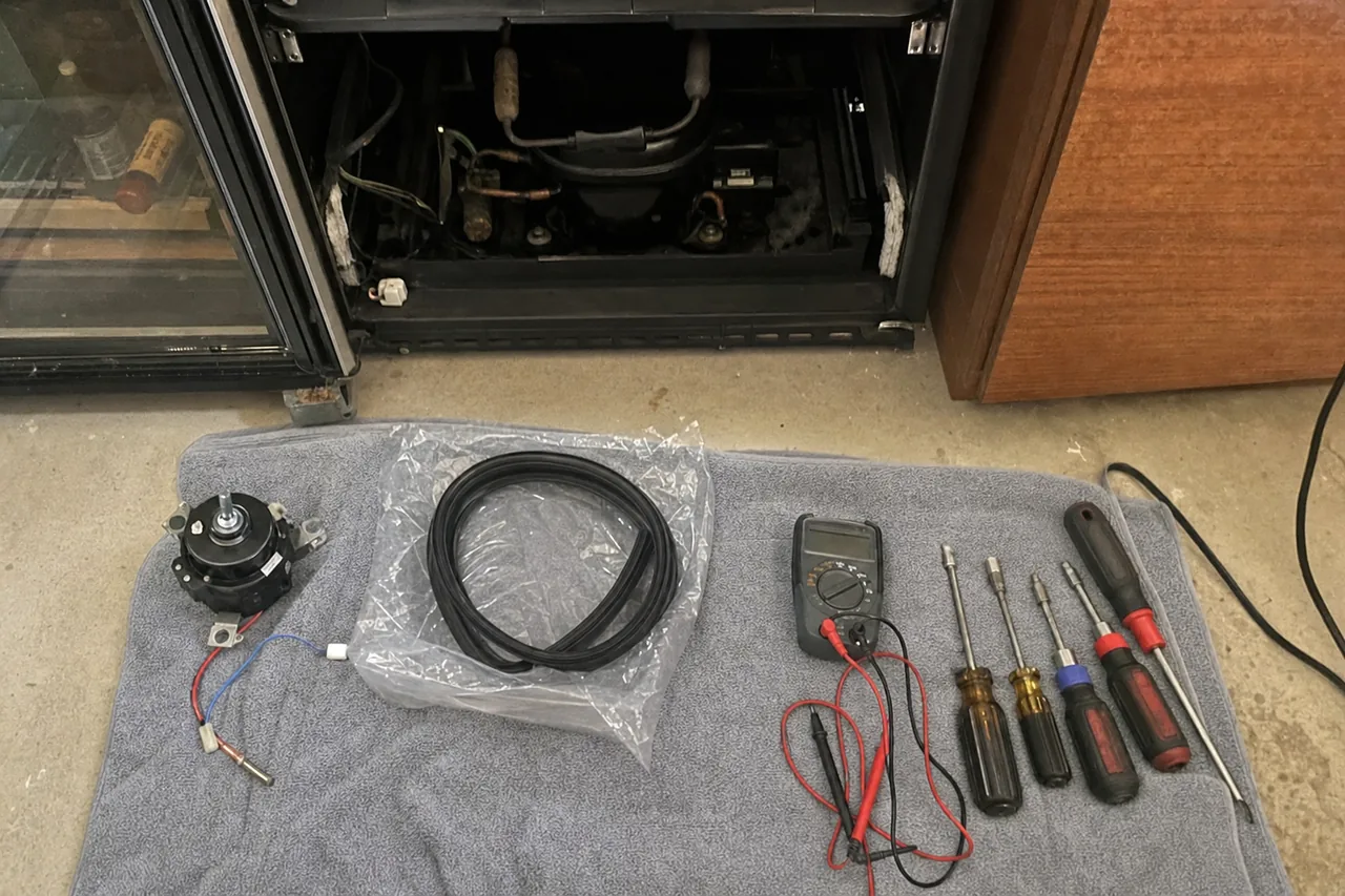 A folded grey towel on a concrete floor holding four nut drivers, two screwdrivers, a multimeter with red and black leads, a bagged replacement door gasket and a condenser fan motor, with an open machine compartment behind.