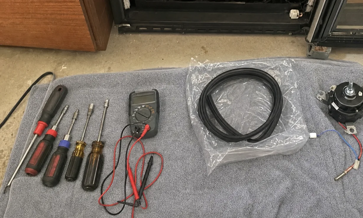 Hand tools and a multimeter laid out in a line on a towel on a bare concrete floor, the power lead of the appliance trailing away to the left.