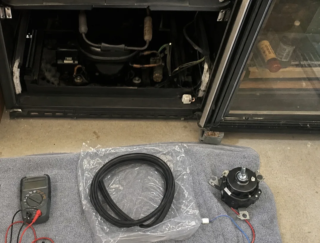 A glass-doored wine cabinet standing beside an opened refrigeration base, bottles lying on their wooden shelves behind the glass and a replacement gasket in a bag on the floor.