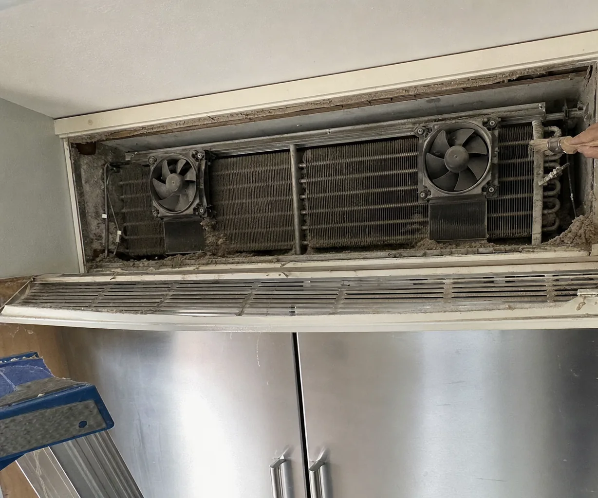The left half of an opened machine compartment above a built-in refrigerator: one condenser fan, a bank of matted coil fins, and wiring running back into the cabinet wall.