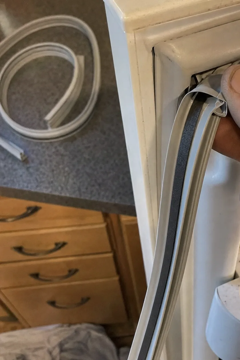A replacement door gasket coiled on a dark kitchen worktop behind an open refrigerator door, wooden drawer fronts below and a cloth on the floor.
