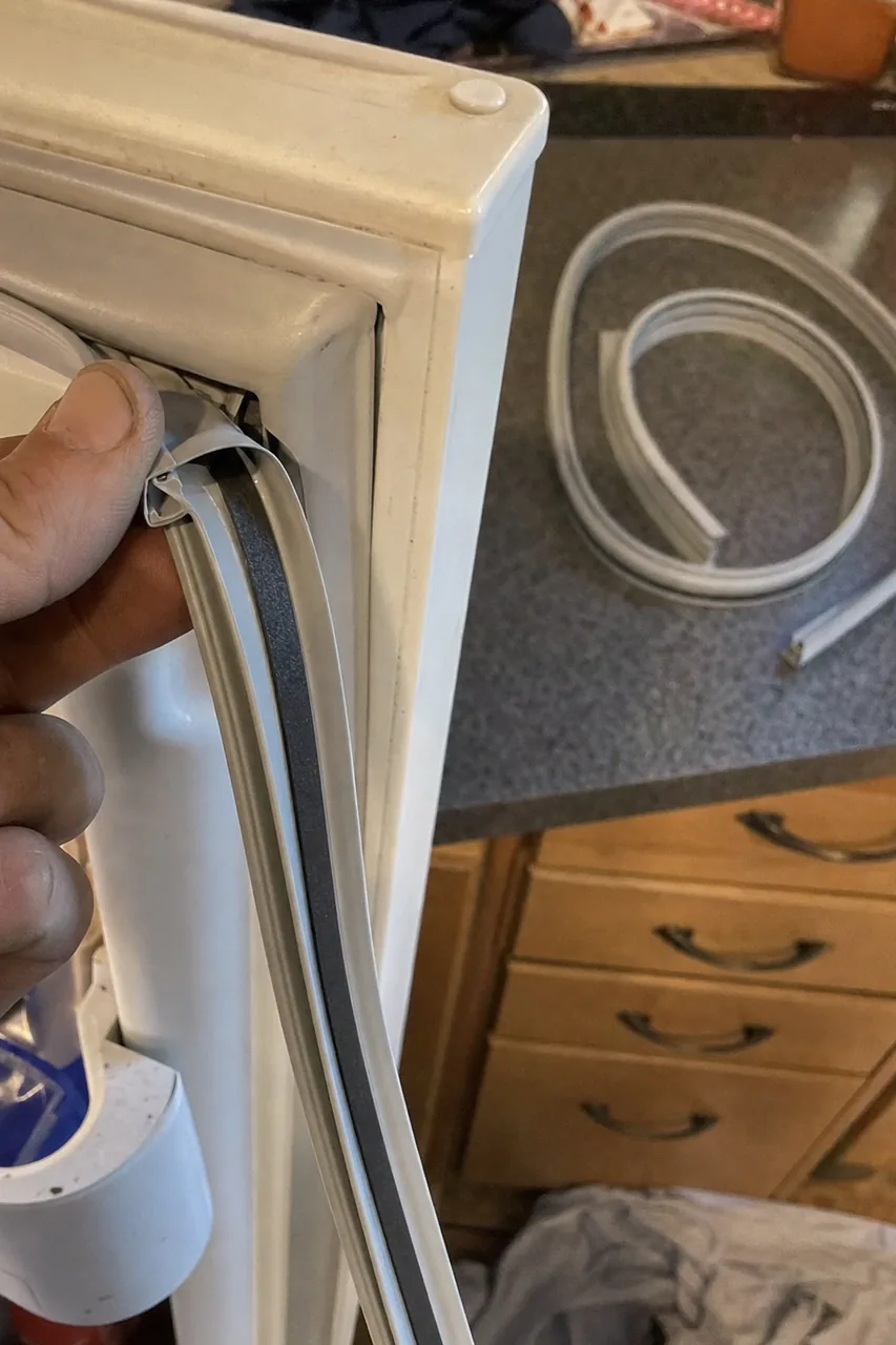 Fingers drawing a used magnetic gasket out of the channel in a refrigerator door frame, the dark sealing face standing proud of the liner, a coiled replacement lying on the worktop behind.