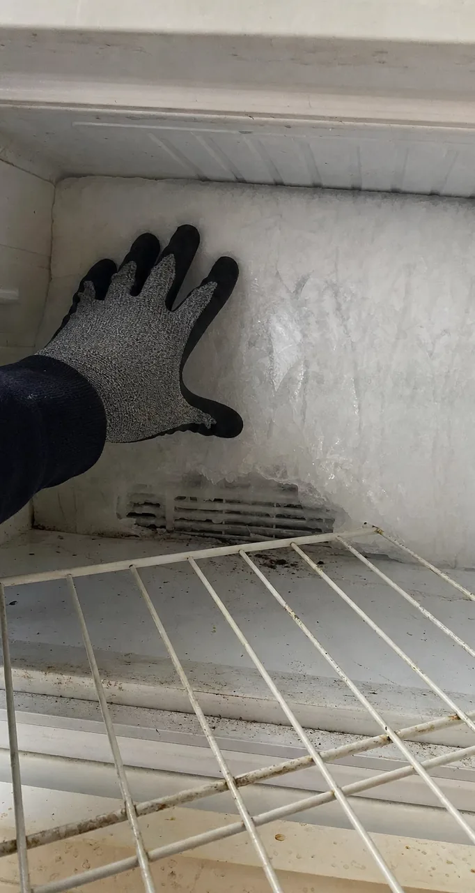Frost sheeted unevenly across the back of a freezer compartment, heavier at the top than the bottom, with the outline of the vent showing through the ice.