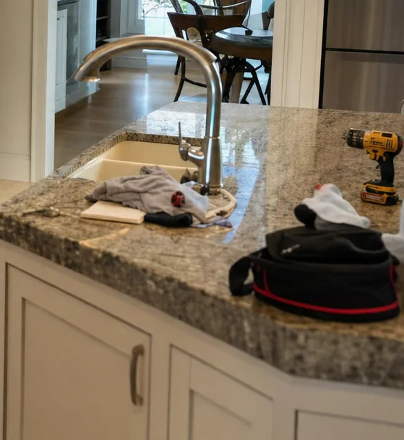 A granite island worktop carrying a cordless drill, a folded rag and an open tool bag, with the kitchen sink and mixer tap behind them.