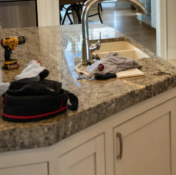 Close on the corner of a granite island where a tool bag, a drill and a cloth have been set down beside the sink, cabinet doors below in painted timber.